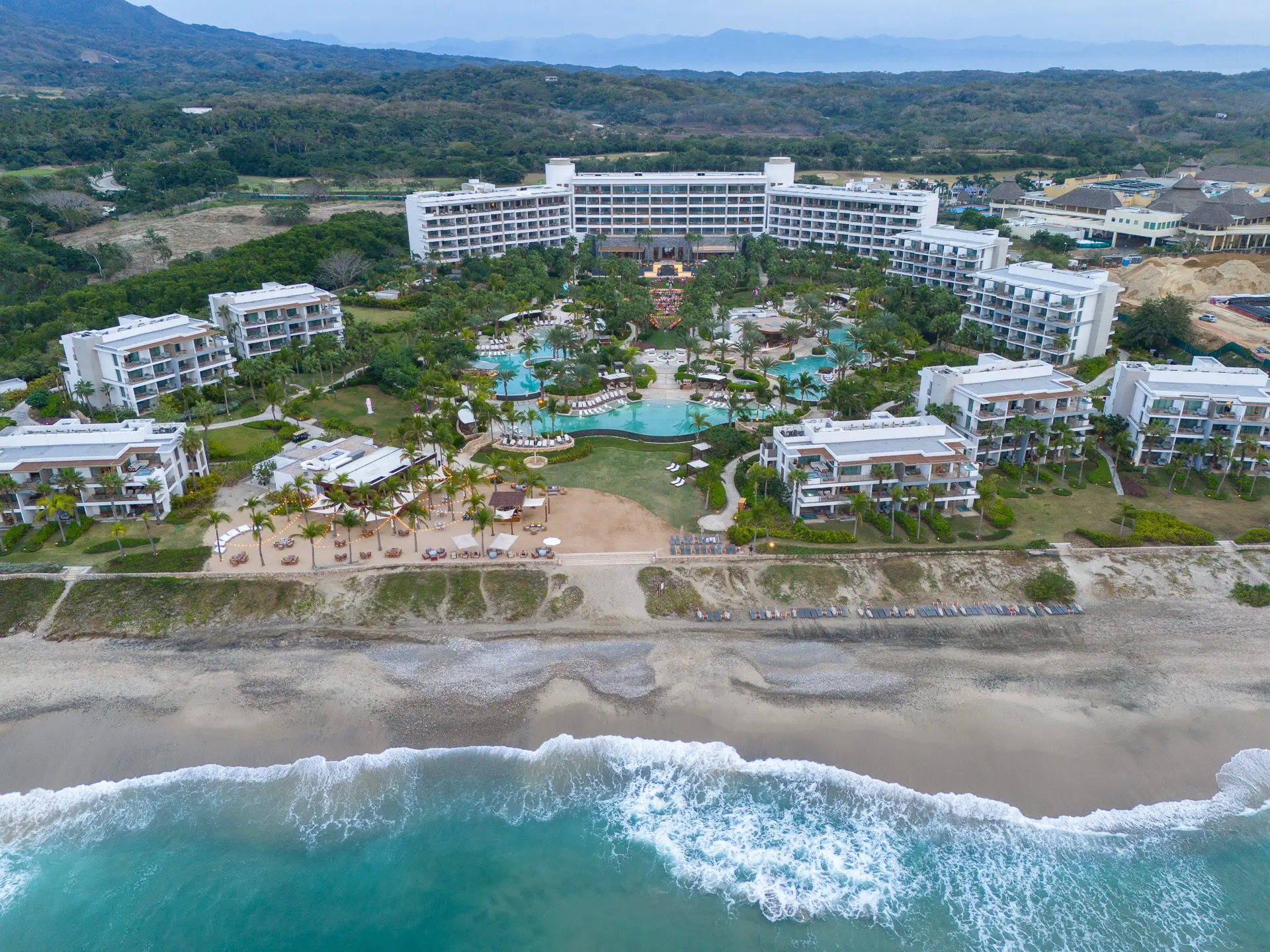 Vista aérea de un resort frente a la playa con piscinas y orilla de arena.