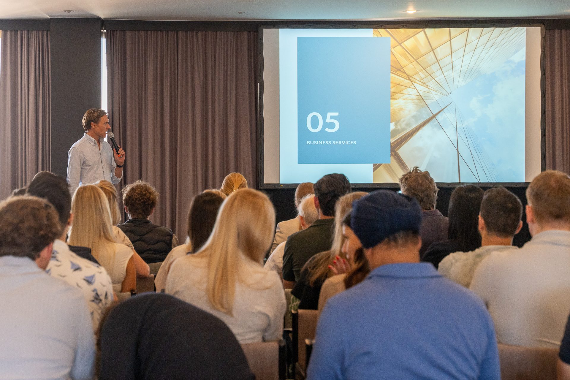 A man giving a presentation in front of a corporate audience, with a screen displaying slides.